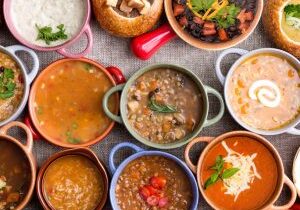 High Angle View of Various Comforting and Savory Gourmet Soups Served in Bread Bowls and Handled Dishes and Topped with Variety of Garnishes on Table Surface with Gray Tablecloth